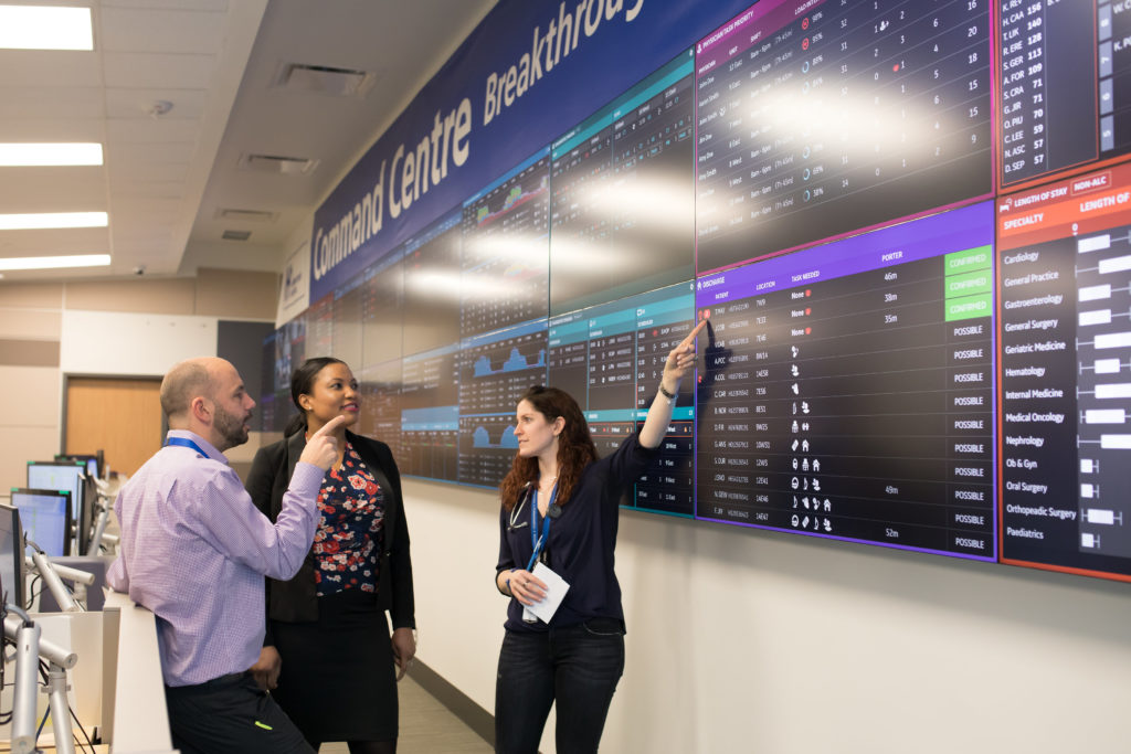 A man and two women working in Humber's Command Centre point to data on the digital tiles. They are looking at each other.