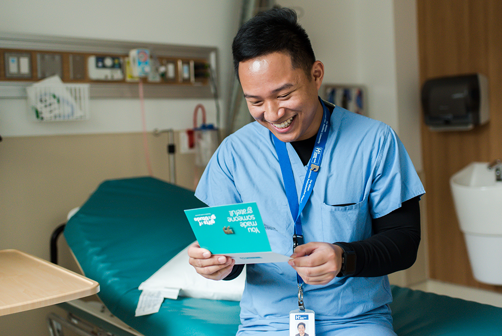 A nurse reads a card that he received from a grateful patient. The card has a Gifts of Gratitude pin on it and says "You made someone grateful" on the front.